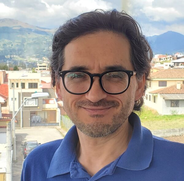 Klever Calle, academic coordinator of react latin america smiles while wearing a blue polo shirt. He has dark wavy hair, glasses, and a short beard. A cityscape with mountains is visible in the background.